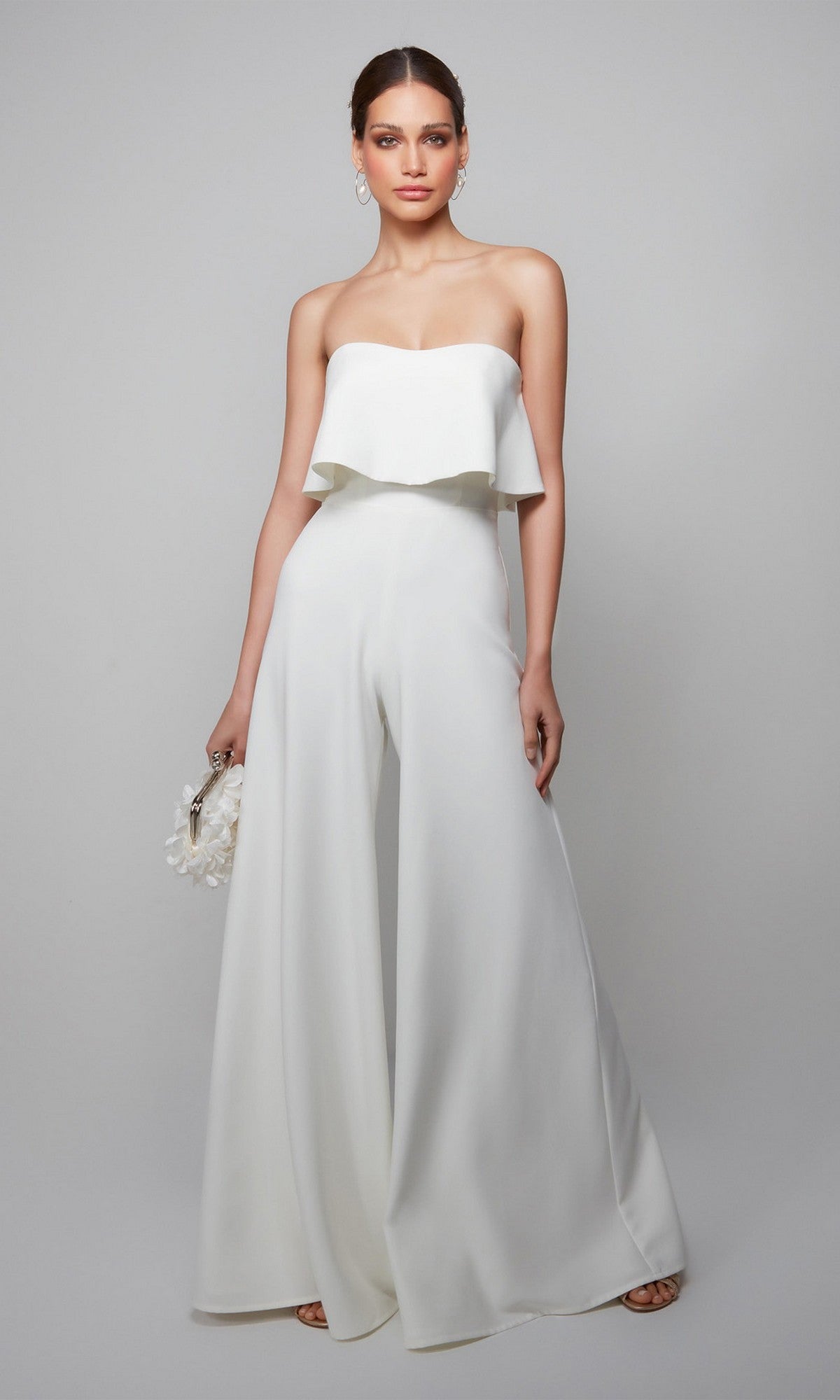 Woman in the Alyce 70015 long formal jumpsuit in ivory, featuring a strapless neckline and wide-leg pants, holding a small bag against a plain gray background.